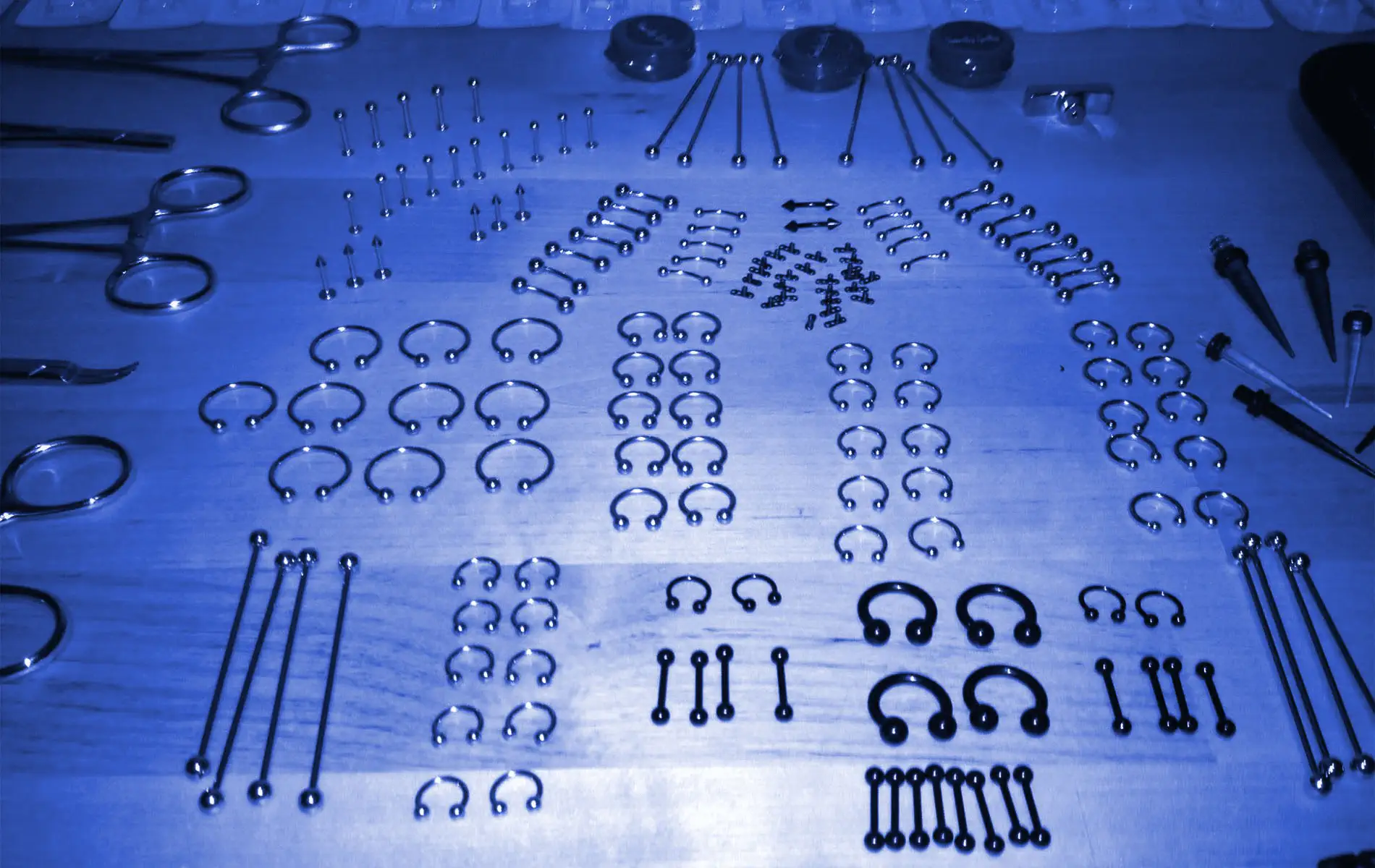 Un lot contenants plusieurs bijoux et pinces pour faire du perçages corporel est étendu sur une table pour servir d'image avec filtre bleu de la page prix body piercing Montréal.