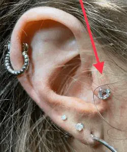 Sur cette oreille ayant plusieurs perçages, une flèche rouge pointe un tragus piercing fait avec bijou en titanium et une pierre blanche de 4mm de grosseur.